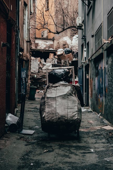 A narrow urban alleyway cluttered with accumulated rubbish, including a large, black, heavy-duty garbage bag in the foreground, partially open revealing dark waste inside, positioned on a damp, worn a