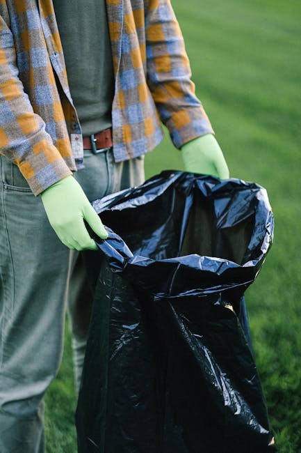 A narrow urban alleyway cluttered with accumulated rubbish, including a large, black, heavy-duty garbage bag in the foreground, partially open revealing dark waste inside, positioned on a damp, worn a