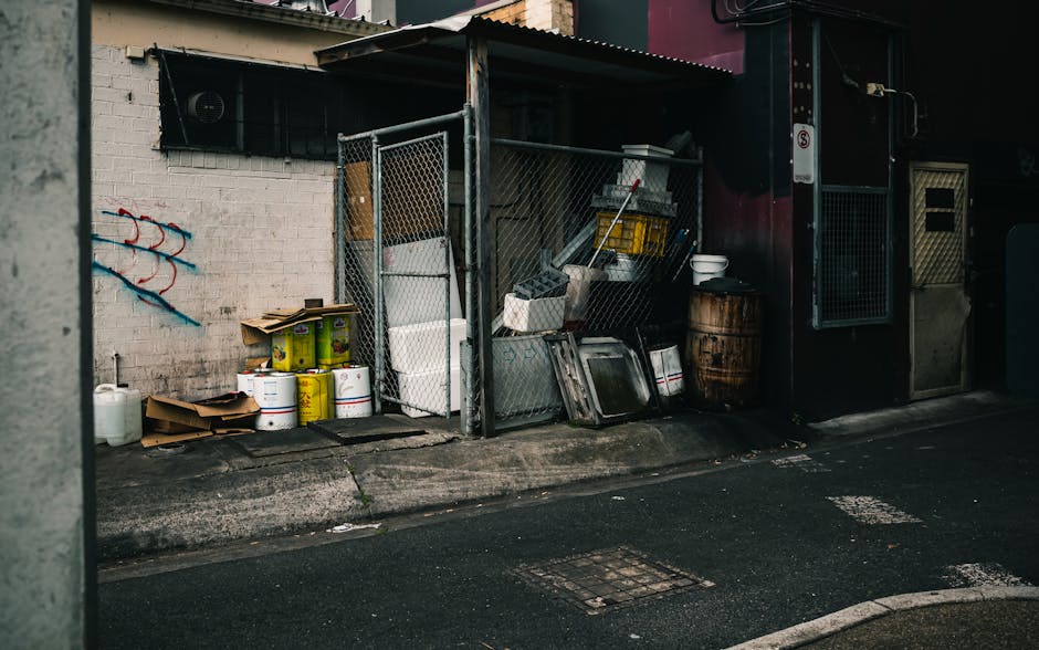 A small outdoor area next to a building features a metal storage shed with a corrugated roof, containing various waste items. In front of the shed, there are several large white plastic buckets with y