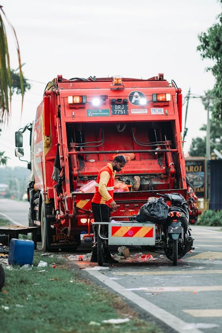 A narrow urban street scene during daylight showing a group of workers in high-visibility vests and hard hats collecting and clearing bulky rubbish near a black waste skip. The garbage appears to incl